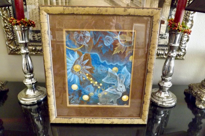 May include: A framed painting of a white rabbit with large ears, surrounded by blue and gold abstract shapes and leaves. The frame is brown with a gold trim.