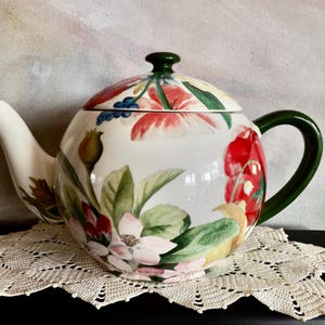 May include: A white ceramic teapot with a floral design featuring red, pink, and green flowers and leaves. The lid and handle are green. The teapot is sitting on a cream-colored lace doily.