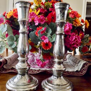 Puede incluir: Dos candelabros plateados de cristal de mercurio con un acabado envejecido. Los candelabros están frente a un colorido arreglo floral con flores rosas, naranjas y amarillas. El arreglo está en un jarrón decorativo.
