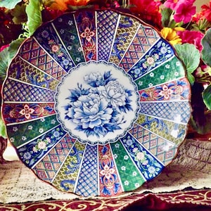 May include: A decorative ceramic plate with a floral design. The plate features a central image of blue flowers surrounded by a patterned border in shades of green, blue, and red. The plate is round and has a scalloped edge.
