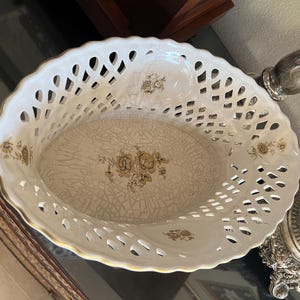 Puede incluir: Una cesta ovalada de cer&aacute;mica con un borde calado de estilo celos&iacute;a en blanco y un borde festoneado dorado. La base interior presenta un acabado de esmalte craquelado color crema decorado con un motivo floral central en tonos marr&oacute;n y oro.