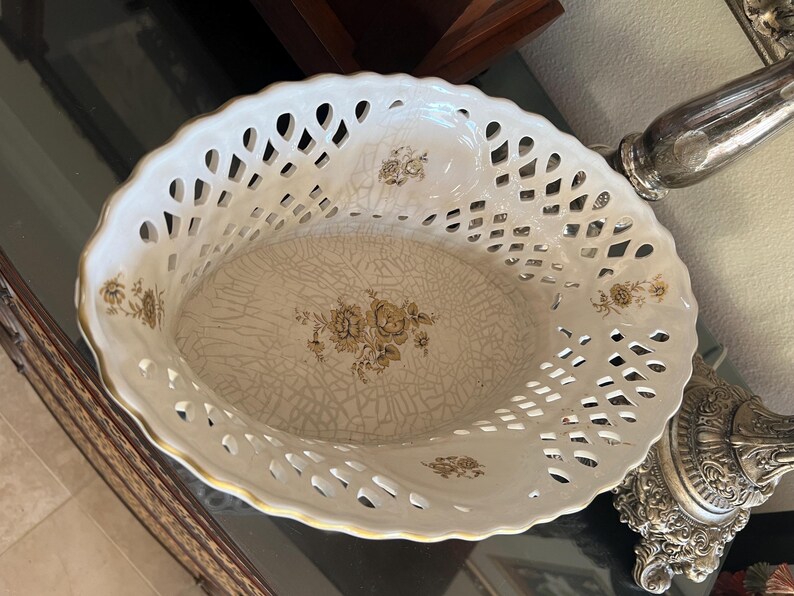 Puede incluir: Una cesta ovalada de cer&aacute;mica con un borde calado de estilo celos&iacute;a en blanco y un borde festoneado dorado. La base interior presenta un acabado de esmalte craquelado color crema decorado con un motivo floral central en tonos marr&oacute;n y oro.