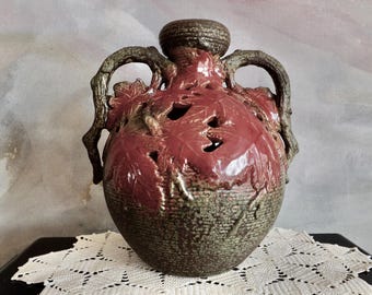 Puede incluir: Un jarrón de cerámica con un diseño de hojas marrón rojizo y dos asas arqueadas. El jarrón tiene una base texturizada de tejido de cesta y una parte superior marrón oscuro. Las asas son de un marrón más claro, parecidas a ramas.