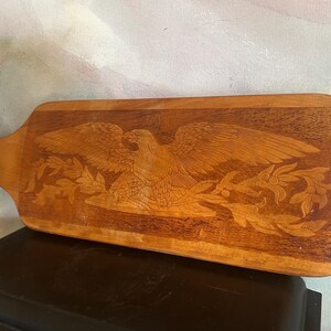 May include: A rectangular wooden serving board with a handle featuring a light-colored leather cord loop. The surface displays a detailed, laser-engraved design of a spread-winged eagle perched above a branch with leaves and berries.