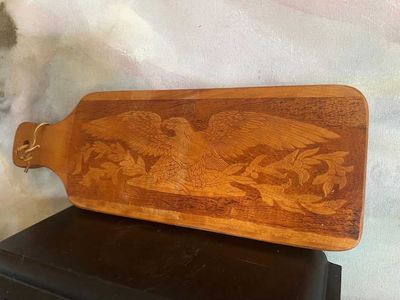 May include: A rectangular wooden serving board with a handle featuring a light-colored leather cord loop. The surface displays a detailed, laser-engraved design of a spread-winged eagle perched above a branch with leaves and berries.