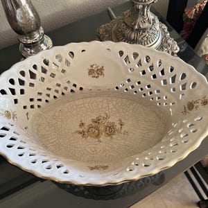 Puede incluir: Un cuenco ovalado de cer&aacute;mica con un borde calado de estilo celos&iacute;a en blanco y un acabado dorado. La base interior muestra un patr&oacute;n floral marr&oacute;n sobre un esmalte craquelado, con motivos florales adicionales en los lados del cuenco.