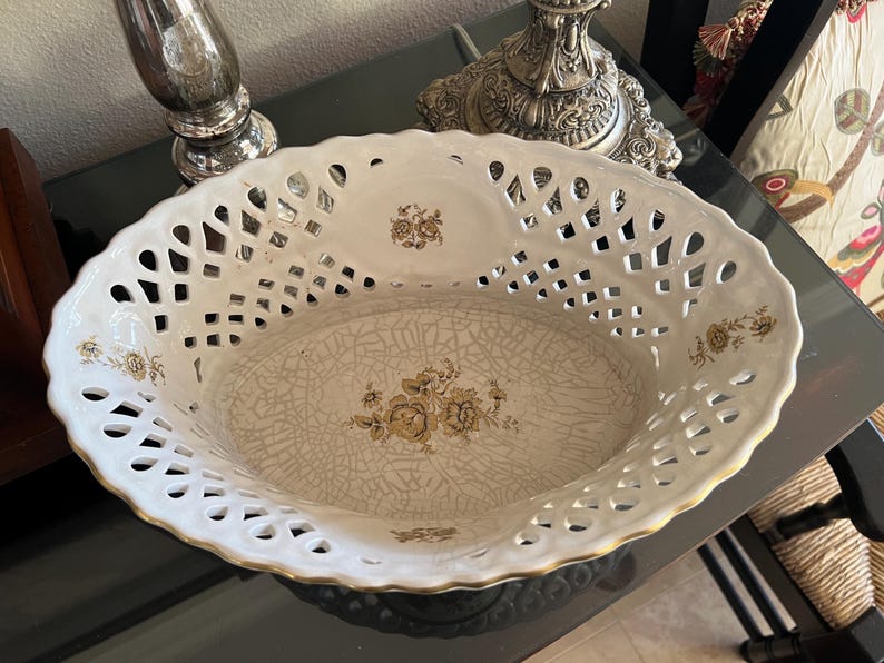 Puede incluir: Un cuenco ovalado de cer&aacute;mica con un borde calado de estilo celos&iacute;a en blanco y un acabado dorado. La base interior muestra un patr&oacute;n floral marr&oacute;n sobre un esmalte craquelado, con motivos florales adicionales en los lados del cuenco.