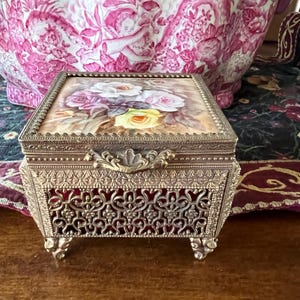 May include: A rectangular metal jewelry box with an ornate filigree base and four decorative feet. The lid features a painted floral design with white, pink, and yellow roses. The box has a textured metal frame and a central latch handle.