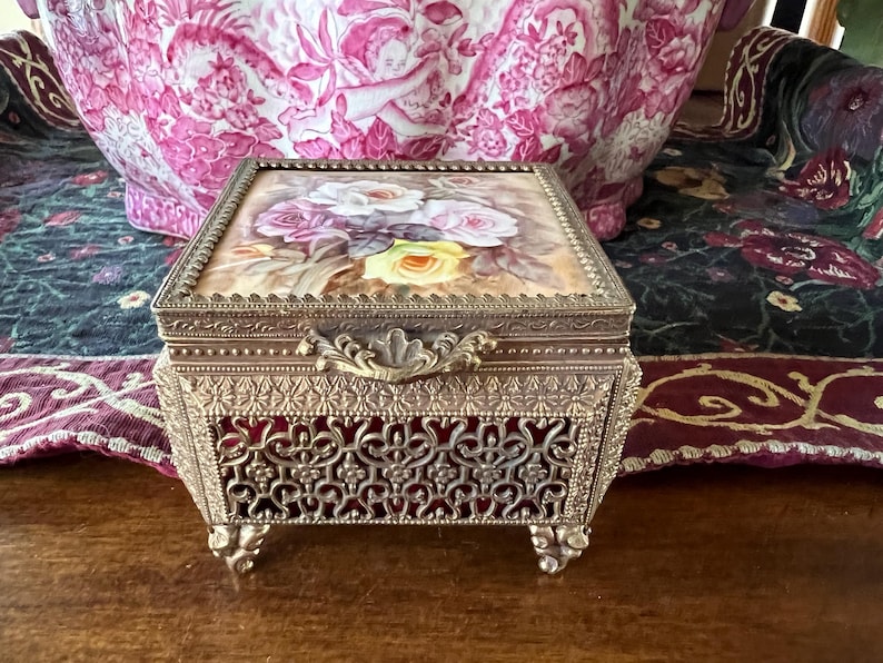 May include: A rectangular metal jewelry box with an ornate filigree base and four decorative feet. The lid features a painted floral design with white, pink, and yellow roses. The box has a textured metal frame and a central latch handle.