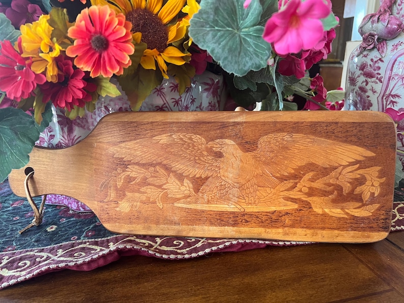 May include: A rectangular wooden cutting board with a rounded handle featuring a hole and a leather cord. The surface displays a detailed wood-burned illustration of a bald eagle with wings spread wide, perched above a branch with leaves.