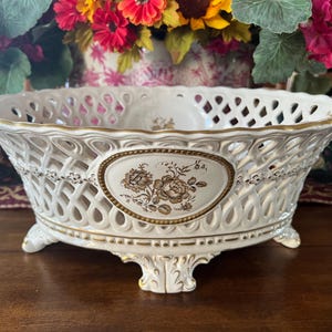 May include: An oval ceramic bowl featuring an intricate lattice-work design with gold-trimmed edges. The center of the bowl displays a detailed brown floral transfer print on an oval medallion. It stands on four ornate, gold-accented feet.