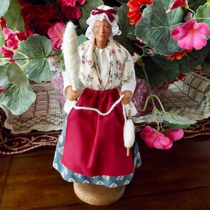 May include: A handcrafted figurine of a woman dressed in traditional attire, holding a spindle and a ball of yarn. The figure wears a white bonnet, a floral-patterned blouse, and a red skirt. The base is a light brown color.