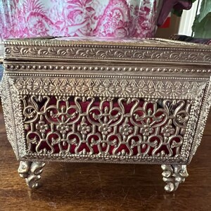 May include: A rectangular metal jewelry box with a silver-toned finish, featuring an intricate filigree pattern on the sides and lid. The box is lined with a deep red velvet fabric visible through the metal cutouts and stands on four ornate feet.