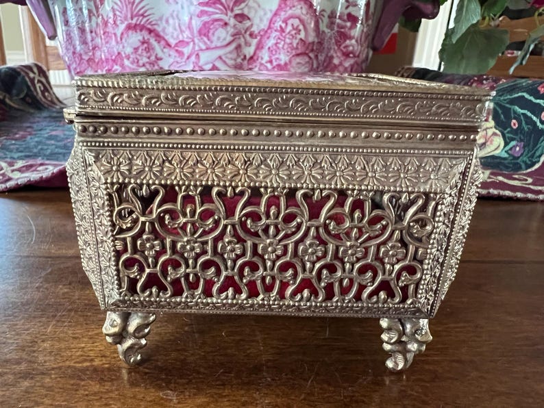May include: A rectangular metal jewelry box with a silver-toned finish, featuring an intricate filigree pattern on the sides and lid. The box is lined with a deep red velvet fabric visible through the metal cutouts and stands on four ornate feet.