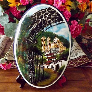 May include: Oval ceramic decorative plate with a hand-painted landscape scene. The artwork depicts a European village with a church, houses, and a gated entrance. The plate is supported by a wooden stand and surrounded by flowers.