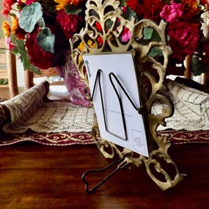 May include: Ornate gold-colored metal picture frame with a black wire card holder. A white card is held in the frame. A colorful bouquet of flowers is in the background. The frame is on a dark wooden surface.