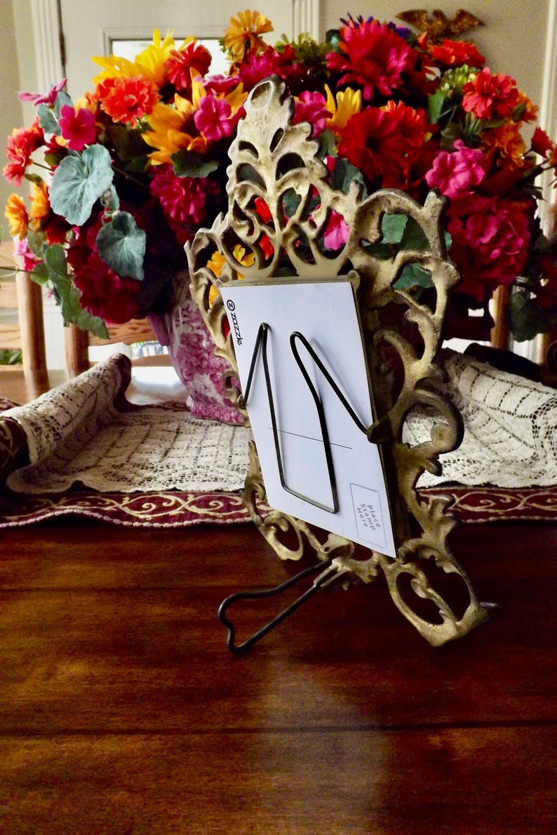 May include: Ornate gold-colored metal picture frame with a black wire card holder. A white card is held in the frame. A colorful bouquet of flowers is in the background. The frame is on a dark wooden surface.