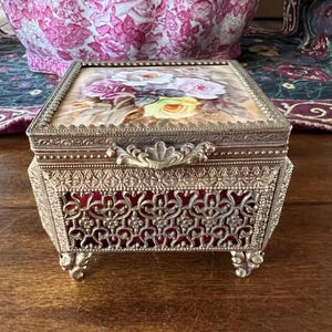 May include: A rectangular metal jewelry box with an ornate filigree design and four decorative feet. The lid features a painted floral scene with white, pink, and yellow roses. The sides of the box reveal a deep red velvet lining behind the metal.