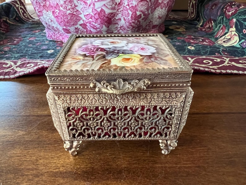 May include: A rectangular metal jewelry box with an ornate filigree design and four decorative feet. The lid features a painted floral scene with white, pink, and yellow roses. The sides of the box reveal a deep red velvet lining behind the metal.