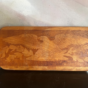 May include: A rectangular wooden serving board with a handle on the left side. The surface features a detailed laser-engraved design of a bald eagle with wings spread wide, perched above a decorative arrangement of leafy branches and berries.