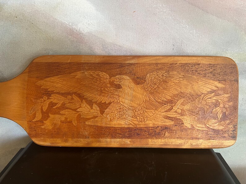 May include: A rectangular wooden serving board with a handle on the left side. The surface features a detailed laser-engraved design of a bald eagle with wings spread wide, perched above a decorative arrangement of leafy branches and berries.