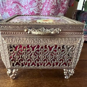 May include: A rectangular metal jewelry box with an ornate gold-tone filigree frame and four decorative feet. The box features a hinged lid with a floral print insert and a deep red velvet lining visible through the intricate metal lattice sides.