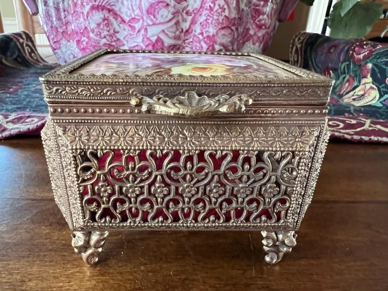 May include: A rectangular metal jewelry box with an ornate gold-tone filigree frame and four decorative feet. The box features a hinged lid with a floral print insert and a deep red velvet lining visible through the intricate metal lattice sides.