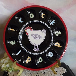 May include: A black ceramic egg holder with a red rim. The center features a white hen illustration. The holder has 12 egg-shaped indentations with colorful letters spelling "cock a doodle poo".