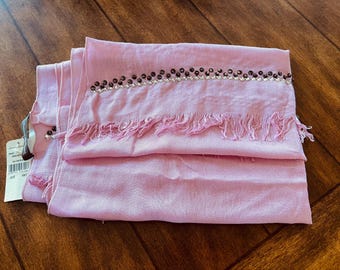 Puede incluir: Una bufanda rectangular de color rosa claro con una banda horizontal decorativa de pequeñas cuentas de color marrón oscuro y transparentes. La bufanda tiene bordes deshilachados en el dobladillo inferior y una etiqueta de papel blanca.