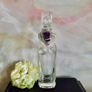 Puede incluir: Una botella de vidrio transparente con un diseño de alambre plateado y un tapón de cristal de amatista púrpura.