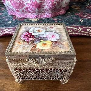 May include: A square metal jewelry box with an ornate filigree pattern on the sides and a hinged lid. The lid features a painted floral design with white, pink, and yellow roses on a beige background. The box has a decorative metal latch.