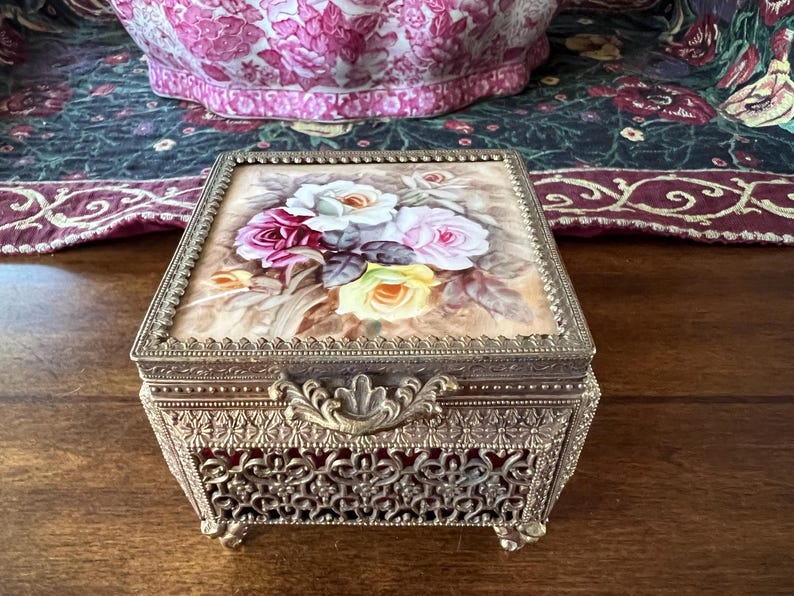 May include: A square metal jewelry box with an ornate filigree pattern on the sides and a hinged lid. The lid features a painted floral design with white, pink, and yellow roses on a beige background. The box has a decorative metal latch.