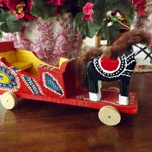 Puede incluir: Un carro de juguete de madera rojo con un caballo blanco y negro tirando de &eacute;l. El carro est&aacute; decorado con flores y remolinos coloridos. El caballo tiene una melena y una cola marrones.