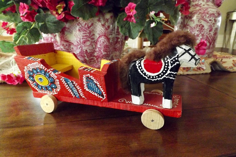 Puede incluir: Un carro de juguete de madera rojo con un caballo blanco y negro tirando de &eacute;l. El carro est&aacute; decorado con flores y remolinos coloridos. El caballo tiene una melena y una cola marrones.