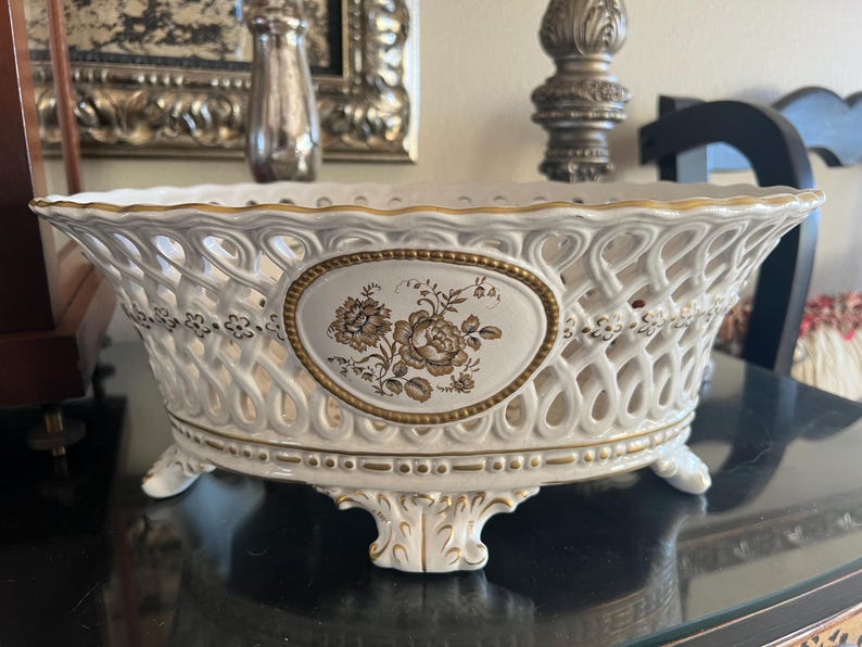 Puede incluir: Un cuenco ovalado de cer&aacute;mica con un intrincado dise&ntilde;o de celos&iacute;a en los laterales, un borde festoneado y cuatro pies ornamentados. El centro del cuenco muestra un medall&oacute;n ovalado con una ilustraci&oacute;n floral marr&oacute;n y bordes dorados.