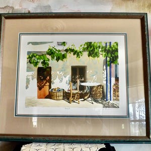 May include: A framed watercolor painting depicting a quaint outdoor scene with a table and chairs set beneath a vine-covered trellis. The painting features a white building with a wooden door and a window, and a basket of fruit sits on the ground.