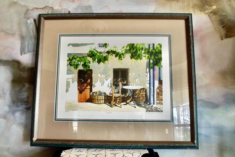 May include: A framed watercolor painting depicting a quaint outdoor scene with a table and chairs set beneath a vine-covered trellis. The painting features a white building with a wooden door and a window, and a basket of fruit sits on the ground.