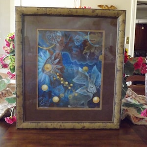 May include: A framed art print with a blue and gold background featuring a white rabbit with large ears and a celestial theme. The print is surrounded by a brown mat and a gold frame.
