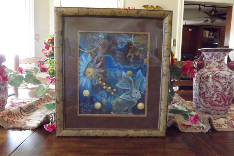 May include: A framed art print with a blue and gold background featuring a white rabbit with large ears and a celestial theme. The print is surrounded by a brown mat and a gold frame.
