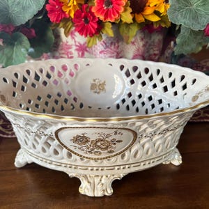 Puede incluir: Un cuenco ovalado de porcelana con cuerpo de celos&iacute;a blanca y bordes dorados. El exterior muestra un panel triangular central con un estampado floral marr&oacute;n. El cuenco se apoya sobre cuatro patas ornamentadas con detalles dorados.