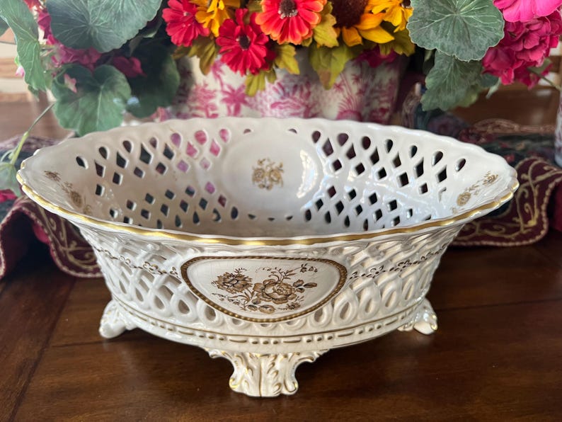 Puede incluir: Un cuenco ovalado de porcelana con cuerpo de celos&iacute;a blanca y bordes dorados. El exterior muestra un panel triangular central con un estampado floral marr&oacute;n. El cuenco se apoya sobre cuatro patas ornamentadas con detalles dorados.