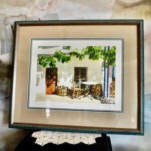 May include: A framed watercolor painting depicting a courtyard scene with a table and chairs, surrounded by greenery and a stone building. The painting is in a green frame with a white mat.