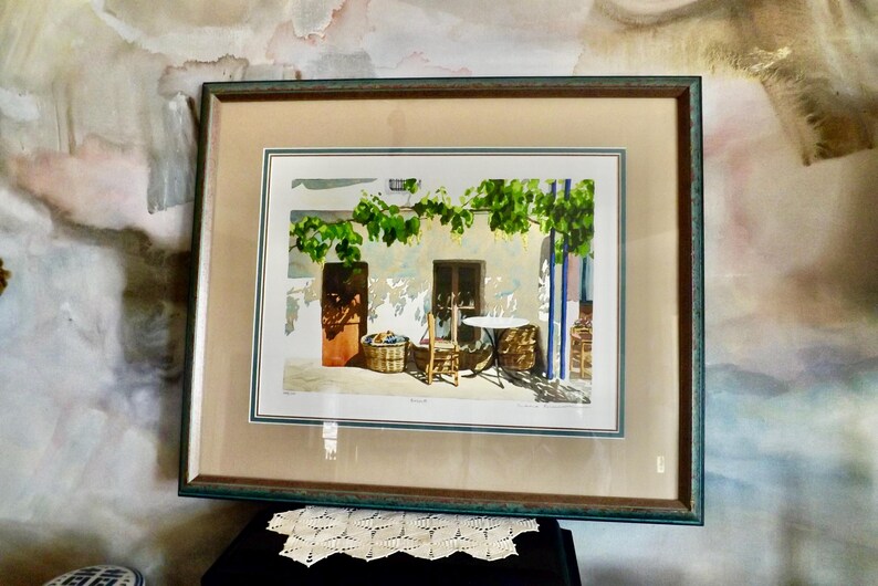 May include: A framed watercolor painting depicting a courtyard scene with a table and chairs, surrounded by greenery and a stone building. The painting is in a green frame with a white mat.