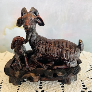 May include: A dark brown carved wooden sculpture depicting a mother goat and a small kid resting together. The figures are mounted on a decorative, carved wooden base featuring floral motifs, showcasing detailed textures and natural wood grain.