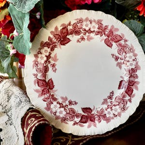 May include: A white ceramic plate with a scalloped edge, adorned with a burgundy floral and leaf pattern. The design encircles the plate's rim, creating a decorative border. The plate is set against a backdrop of colorful flowers.