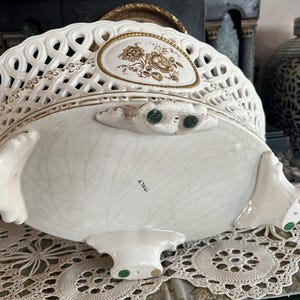 Puede incluir: Una cesta ovalada de cer&aacute;mica con cuerpo de rejilla color crema, con medallones florales con bordes dorados y cuatro patas ornamentadas. La parte inferior de la base muestra un acabado craquelado y la palabra ITALY impresa en tinta negra.