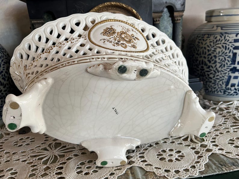 Puede incluir: Una cesta ovalada de cer&aacute;mica con cuerpo de rejilla color crema, con medallones florales con bordes dorados y cuatro patas ornamentadas. La parte inferior de la base muestra un acabado craquelado y la palabra ITALY impresa en tinta negra.