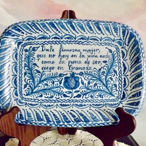 May include: A rectangular ceramic plate with a blue and white floral design and text. The plate is supported by a wooden stand. The text on the plate reads, "Dale limosna mujer, que no hay en la vida nada como la pena de ser, ciego en Granada."