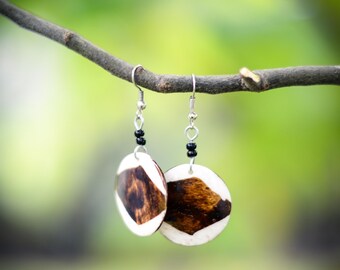 May include: A pair of wooden earrings with a white and brown geometric pattern. The earrings are hanging from a branch against a green background.