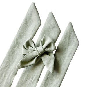 May include: Three sage green linen headbands with a matching bow. The headbands are made of a textured fabric and are arranged diagonally against a white background. The bow is centered on one of the headbands.